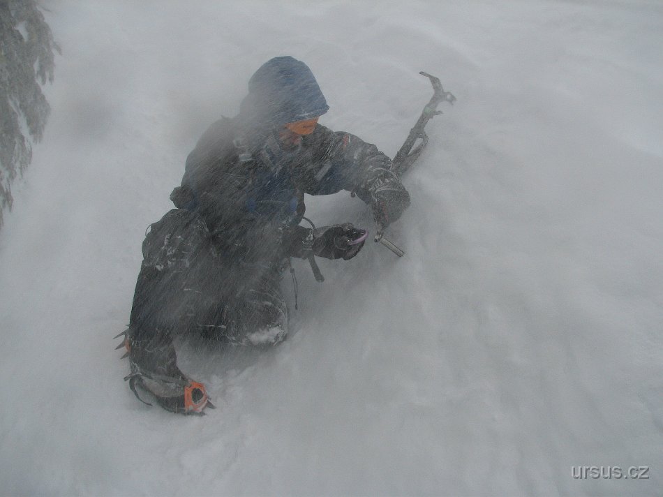 tatry 032.jpg - Došli jsme na chatu a přemýšleli co dál. Venku už nezuřila vichřice, ale orkán, takže nám bylo jasné, že se daleko nedostaneme. Na chatě se nám ale sedět nechtělo, chtěli jsme vystrčit ven aspoň nos. A to se povedlo. Za neustálého strhávání větrem k zemi jsme se "doplazili" asi sto metrů za chatu. Dál to nešlo. Tak jsme si aspoň našli veliký balvan, kolem kterého byl vyfoukaný sníh a byl proto v závětří a trošku si na něm "zablbli". Lukáš právě zkouší zavrtat šroub do ledu.